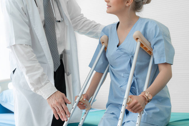 Doctor takes care of patient in crutch at hospital. Physical therapist and leg injury recovery concept. - Photo, Image