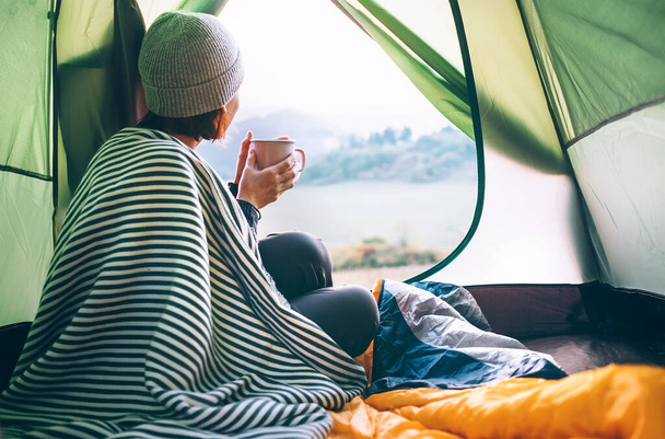 Young woman covered with warm plaid meets cold morning in sitting in a touristic camping tent with a cup of hot tea. Romantic camp traveling concept. - Foto, Imagem