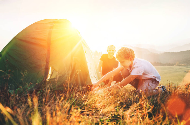 Pai aprende seu filho montar barraca de acampamento na clareira floresta pôr do sol
 - Foto, Imagem