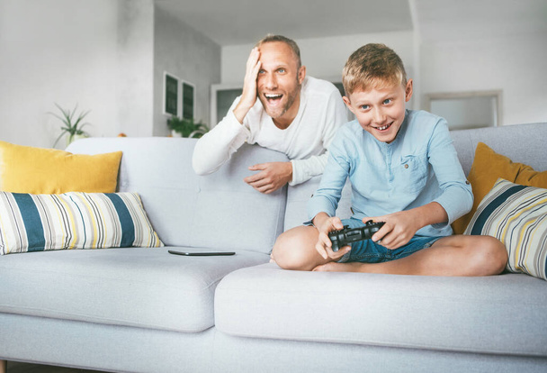 Father watching his son playing TV video game using the gamepad and emotionaly reacts. Liitle kids and playing devices using concept image. - Photo, image