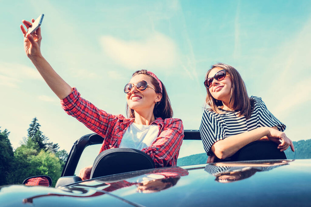 Two female friends take a selfie photo in cabriolet car during their summer voyage - Valokuva, kuva