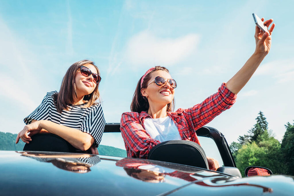 Two female friends take a selfie photo in cabriolet car during their summer voyage - Valokuva, kuva