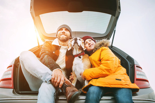 Father and son dressed in yellow warm jacket with beagle dog siting together in car trunk. Long auto journey break. Traveling with pets concept. - Zdjęcie, obraz