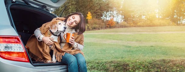 Woman huges her dog with shawls sitting together in car trunk - Foto, Imagen