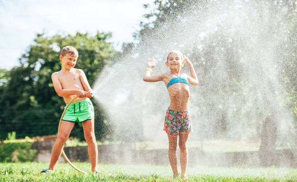 Two happy childs refresh with sprinkling water in hot summer afternoon  - Foto, imagen