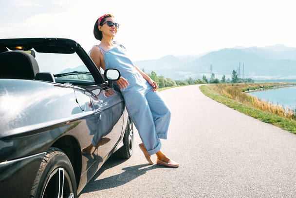 Young woman solo traveler stay near cabriolet car on picturesque mountain road - Foto, imagen