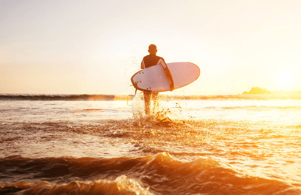 Surfer runs with surfboard towards ocean waves ta sunset time - Фото, зображення