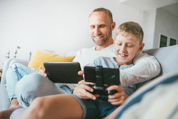 Les joueurs de jeu père et fils s'amusent assis ensemble à la maison sur un canapé confortable, en utilisant la tablette et la manette de jeu - Photo, image