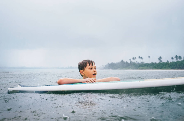 Boy learning to surf under the tropical rain - 写真・画像