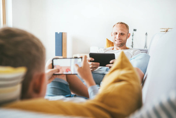  Father and son playing with tablet and gamepad sitting in living room  - Fotó, kép