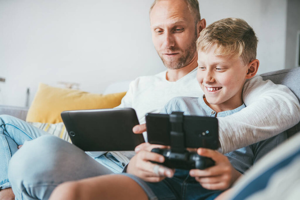 Ojciec i syn gracze gry zabawy siedzieć razem za pomocą tabletu i gamepad w domu na przytulnej sofie - Zdjęcie, obraz