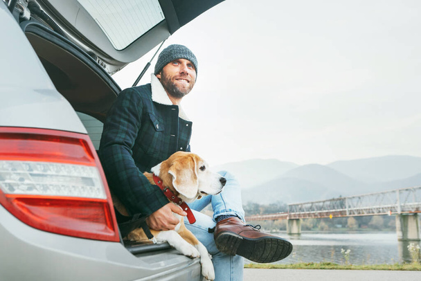Man with beagle dog siting together in car trunk. Late autumn time traveling with pets concept. - Photo, Image