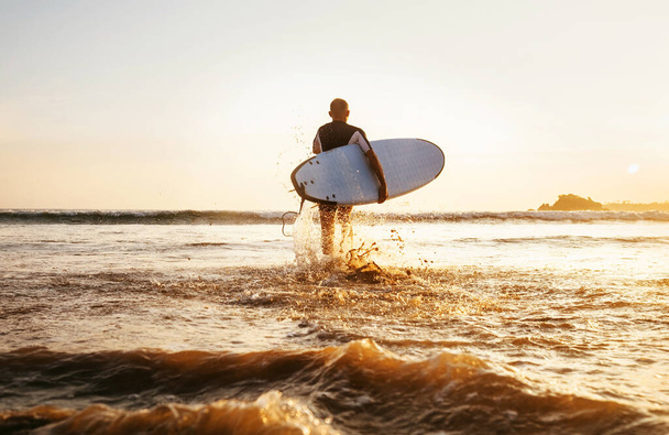 Surfer runs with surfboard towards ocean waves ta sunset time - Foto, Imagen