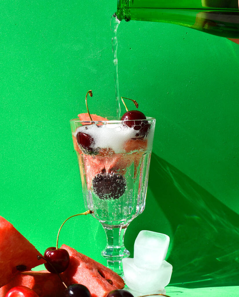 een glas sprankelend water, water stroomt uit de fles. Rond glazen kersen en stukjes watermeloen met ijs - Foto, afbeelding