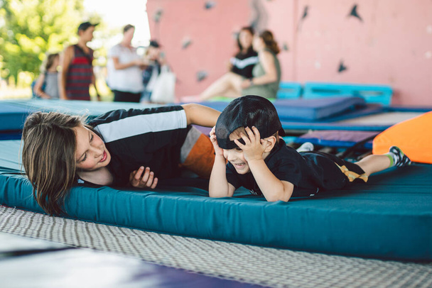 Счастливые дети, играющие на батуте. Братья веселятся, лежа на спортивном коврике на батуте в спортзале снаружи. Мальчики балуются, смеются и обнимаются после тренировки.
. - Фото, изображение