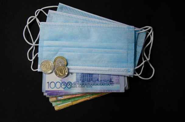 Composition of notes, coins of Kazakhstani tenge and a medical mask on a black background. Kazakh money and a medical mask. - Photo, Image