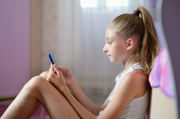 Moderní dívka v bílém tričku sedí a čte si e-knihu na smartphonu. Dívka sedící na podlaze ve svém pokoji - Fotografie, Obrázek