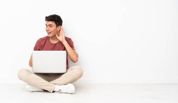 Flor 'da dizüstü bilgisayarıyla oturan genç adam elini kulağına koyarak bir şeyler dinliyor. - Fotoğraf, Görsel