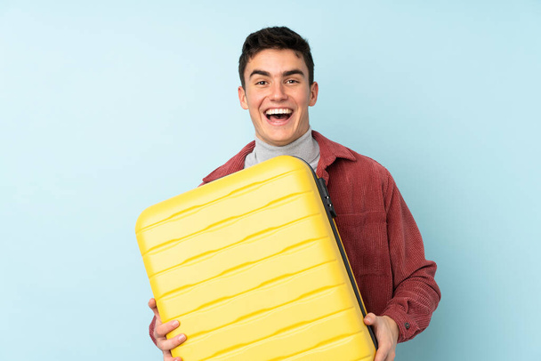 Teenager caucasian man isolated on purple background in vacation with travel suitcase and surprised - Foto, Imagen
