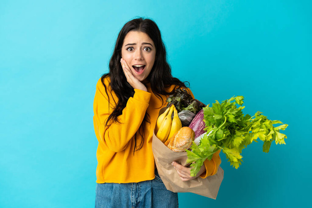 Fiatal nő kezében egy élelmiszer bevásárló táska elszigetelt kék háttér meglepetés és sokkos arckifejezés - Fotó, kép