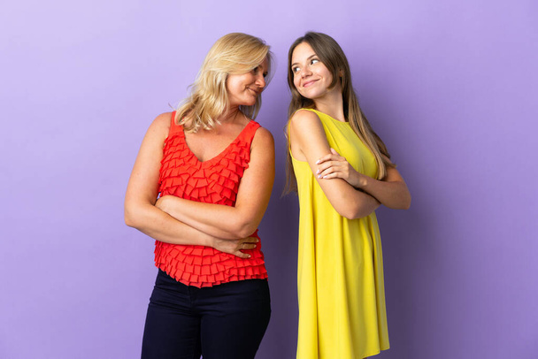 Mom and daughter isolated on purple background looking over the shoulder with a smile - Photo, Image