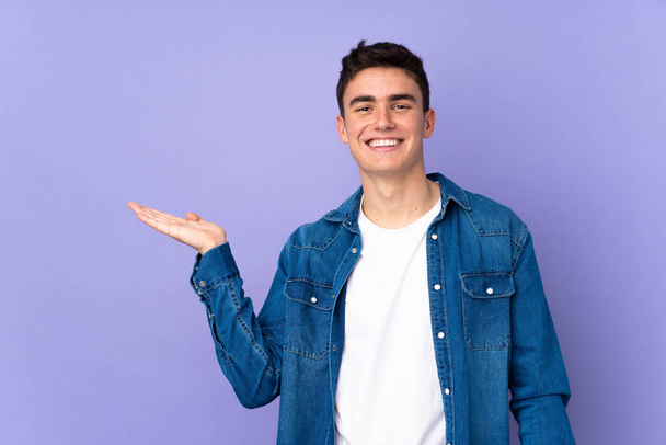 Tiener Kaukasische knappe man geïsoleerd op paarse achtergrond houden copyspace imaginaire op de palm om een advertentie in te voegen - Foto, afbeelding