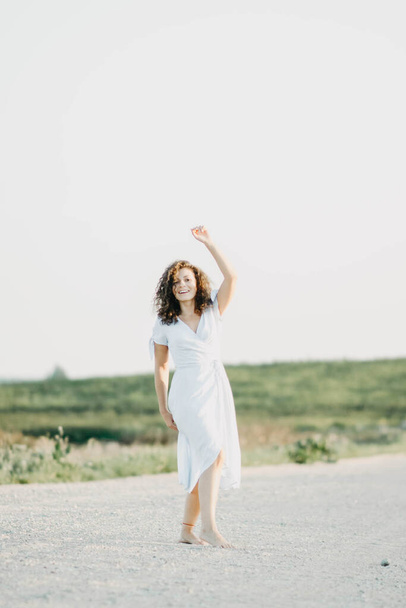 Curly young woman dancing romantically in a blue dress on a sandy road at sunset. - Fotografie, Obrázek
