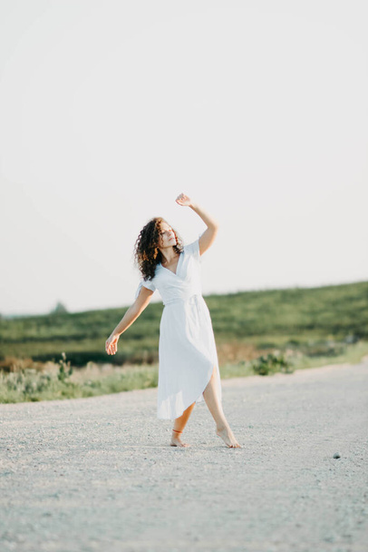 Kudrnatá mladá žena tančí romanticky v modrých šatech na písčité cestě při západu slunce. - Fotografie, Obrázek