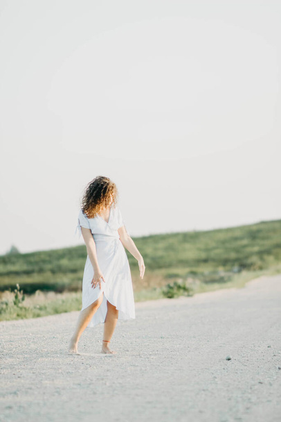 Curly young woman dancing romantically in a blue dress on a sandy road at sunset. - Foto, immagini