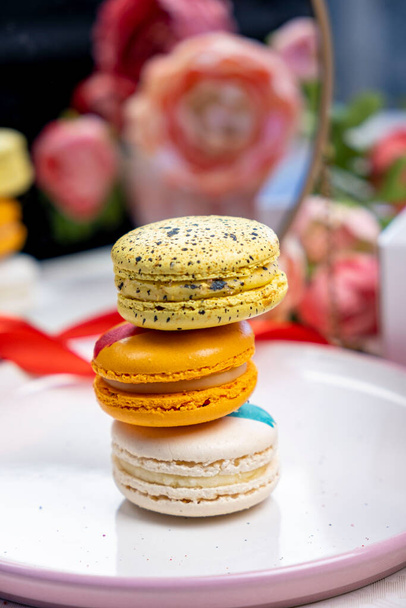 στοίβα από πολύχρωμα μοντέρνα κέικ μπισκότα macaroons στο πιάτο για τη γιορτή ρομαντικά γενέθλια κόμμα, pion λουλούδια, σατέν κορδέλα πάνω όψη, πολλοί διαφορετικοί τύποι γεύσεις των γλυκών macarons διακοσμημένα  - Φωτογραφία, εικόνα