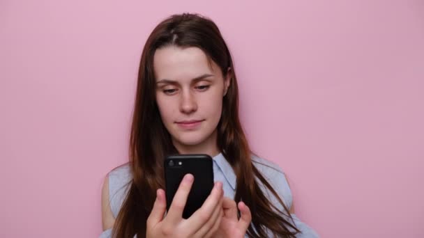 Happy young woman holding smartphone using mobile online apps, smiling brunette girl texting sms message chatting on phone looking at cellphone, wears blue shirt, isolated on pink studio background - Filmagem, Vídeo