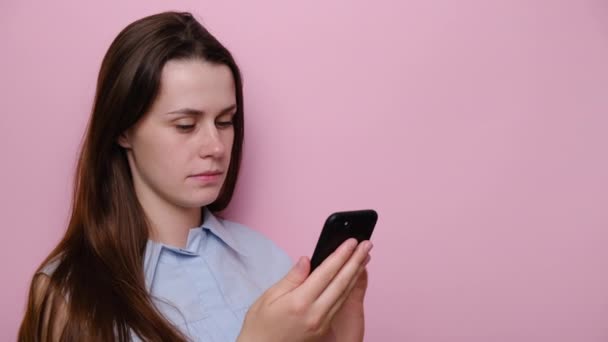 Portrait of sad young girl getting bad news on smartphone, dressed in blue shirt, isolated on pink studio background. Shocked brunette woman feeling bad, worried female checking phone content - Video