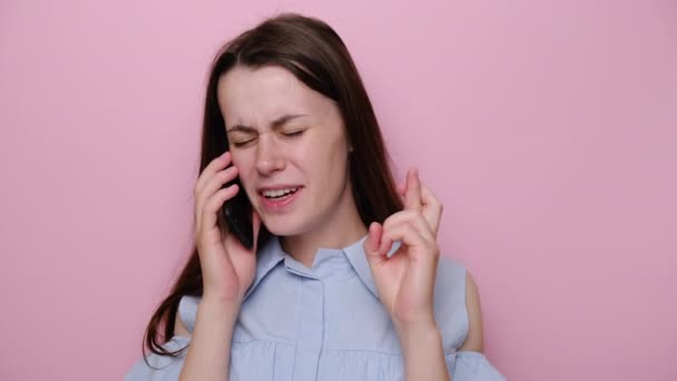 Portrait of cute young woman talking on smartphone, isolated on pink studio background, gesturing finger crossed smiling with hope and eyes closed, dressed in blue shirt. Luck and superstition concept - Felvétel, videó