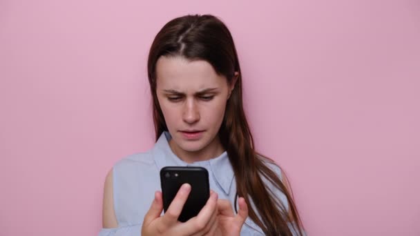 Displeased stressed young woman typing messages on his cell phone, dressed in blue shirt, isolated on pink studio background. Nervous girl thinking over sudden problems after reading sms on smartphone - 映像、動画