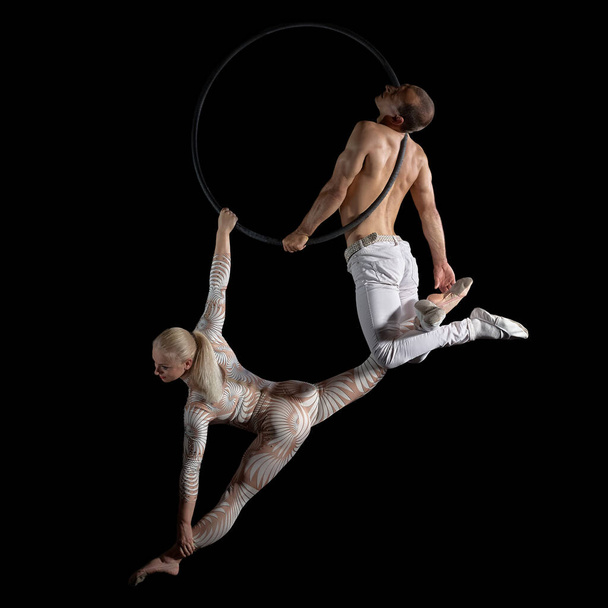A duet of acrobats performed aerial stunts on a suspension ring. Graceful performance of a woman and a man isolated on a black background. - Photo, Image