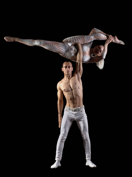 Couple acrobats perform a trick handstand, body balance, isolated on a black background. A girl in a white suit, a guy with a naked muscular torso and white pants. - Zdjęcie, obraz