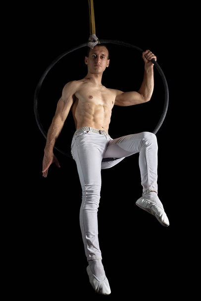 Der Akrobat führt den Trick in einem Ring aus, der in der Luft hängt. Ein Mann mit nacktem muskulösen Oberkörper sitzt in einem Ring in luftiger Höhe. Isoliert auf schwarzem Hintergrund. - Foto, Bild