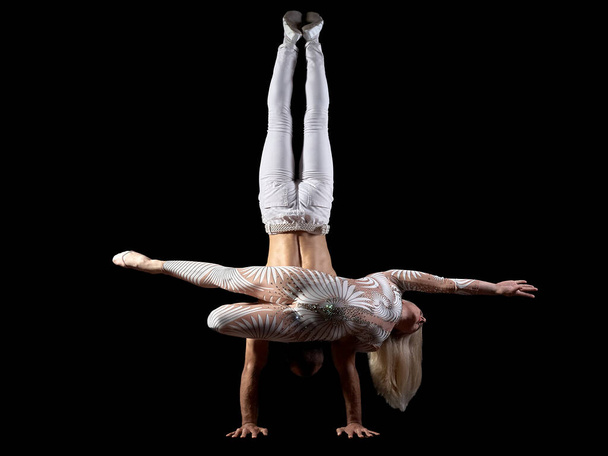 Couple acrobats perform a trick handstand, body balance, isolated on a black background. A girl in a white suit, a guy with a naked muscular torso and white pants. - Photo, Image