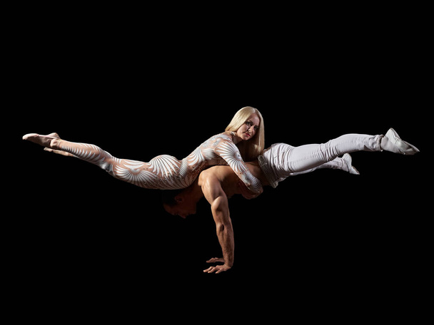 Пара акробатів виконують трюкову сумочку, баланс тіла, ізольовані на чорному тлі. Дівчина в білому костюмі, хлопець з голим м'язистим торсом і білими штанами
. - Фото, зображення