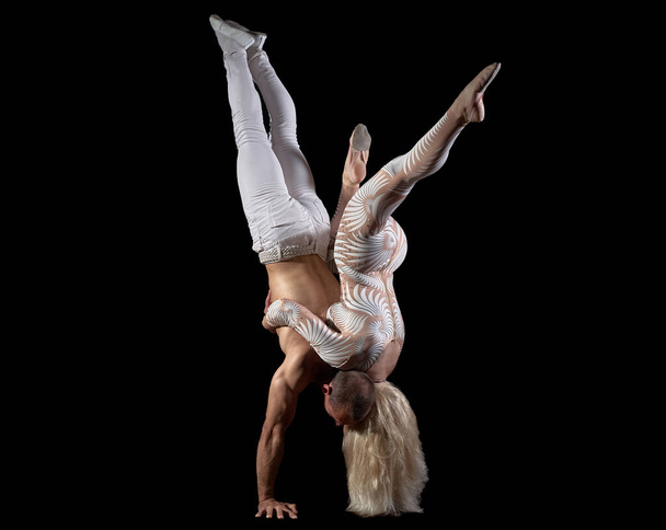 Pár akrobati provést trik stojan, tělo rovnováhu, izolované na černém pozadí. Dívka v bílém obleku, chlap s nahým svalnatým trupem a bílými kalhotami. - Fotografie, Obrázek