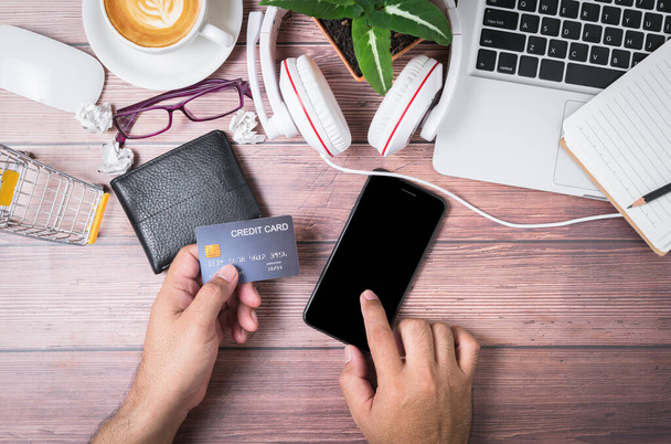 man touch on black screen mobile phone and holding credit card on wallet with laptop on wooden office desk consisting of headphones and hot coffee, shopping online concept - Photo, Image