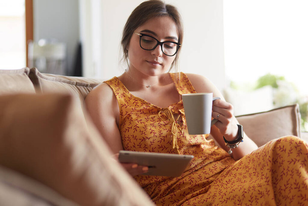 Young woman uses tablet while relaxing at home with coffee on the sofa couch - Foto, Bild