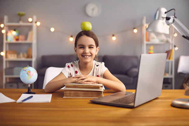 Back to school. Happy schoolgirl with apple ina head smiles while sitting at the table with laptop at home. - Fotografie, Obrázek