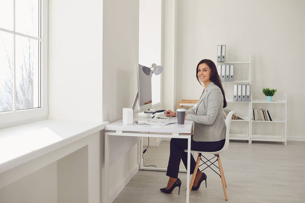 Femme d'affaires souriante dans une veste grise regarde assis sur un lieu de travail à une table dans un bureau blanc. - Photo, image