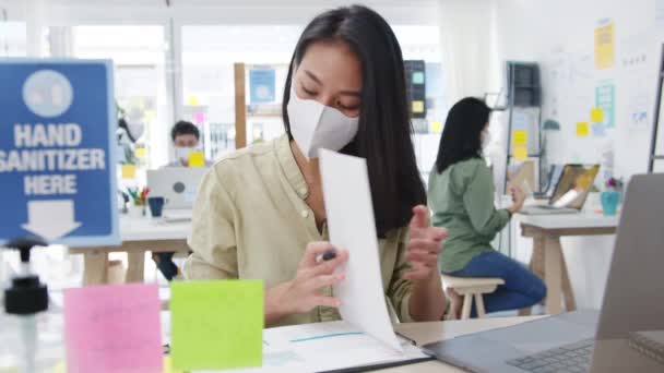 Asiens Unternehmerin mit medizinischer Gesichtsmaske zur sozialen Distanzierung in einer neuen normalen Situation zur Virenprävention, während sie im Büro ihren Laptop wieder bei der Arbeit benutzt. Leben und Arbeit nach dem Coronavirus. - Filmmaterial, Video