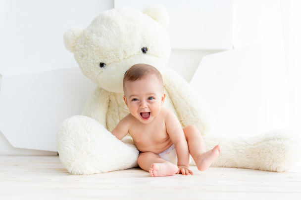 Małe dziecko dziewczyna 6 miesięcy siedzi z dużym miękkim niedźwiedziem w jasnym mieszkaniu w pieluchach. - Zdjęcie, obraz