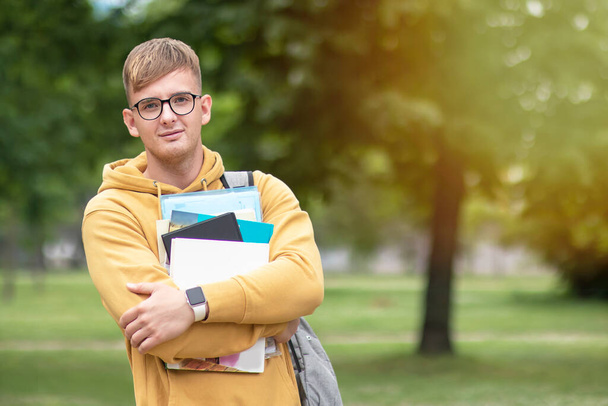 Handsome positive university or college student standing outdoors in park in glasses, holding books, textbooks in his hands, smiling. Cheerful guy young man looking at camera. Education, study concept - Zdjęcie, obraz