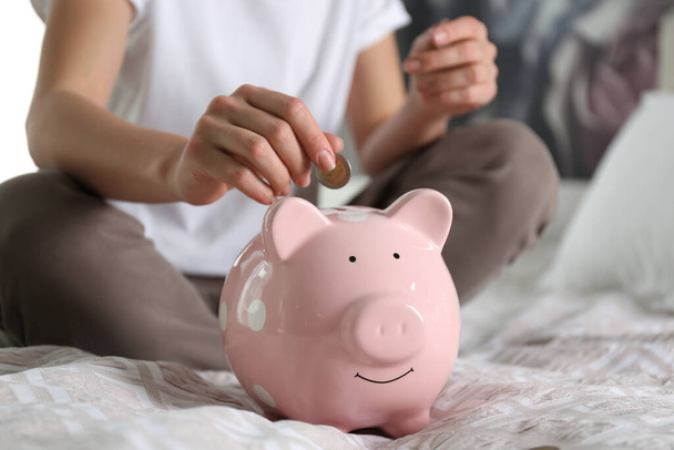 Woman putting money into piggy bank on bed at home, closeup - 写真・画像