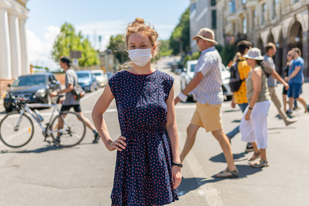 Une jeune jolie fille masquée est debout près d'une foule dans une rue traversant la route, beaucoup de gens marchant masques et ne gardent pas de distance sociale - Photo, image