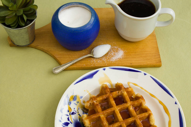 Waffle on a white plate with blue details, with a wooden tray where there is a blue pot of sugar, with a silver spoon, a green plant, and a white pot with caramel sauce, on a green background.. - Photo, Image